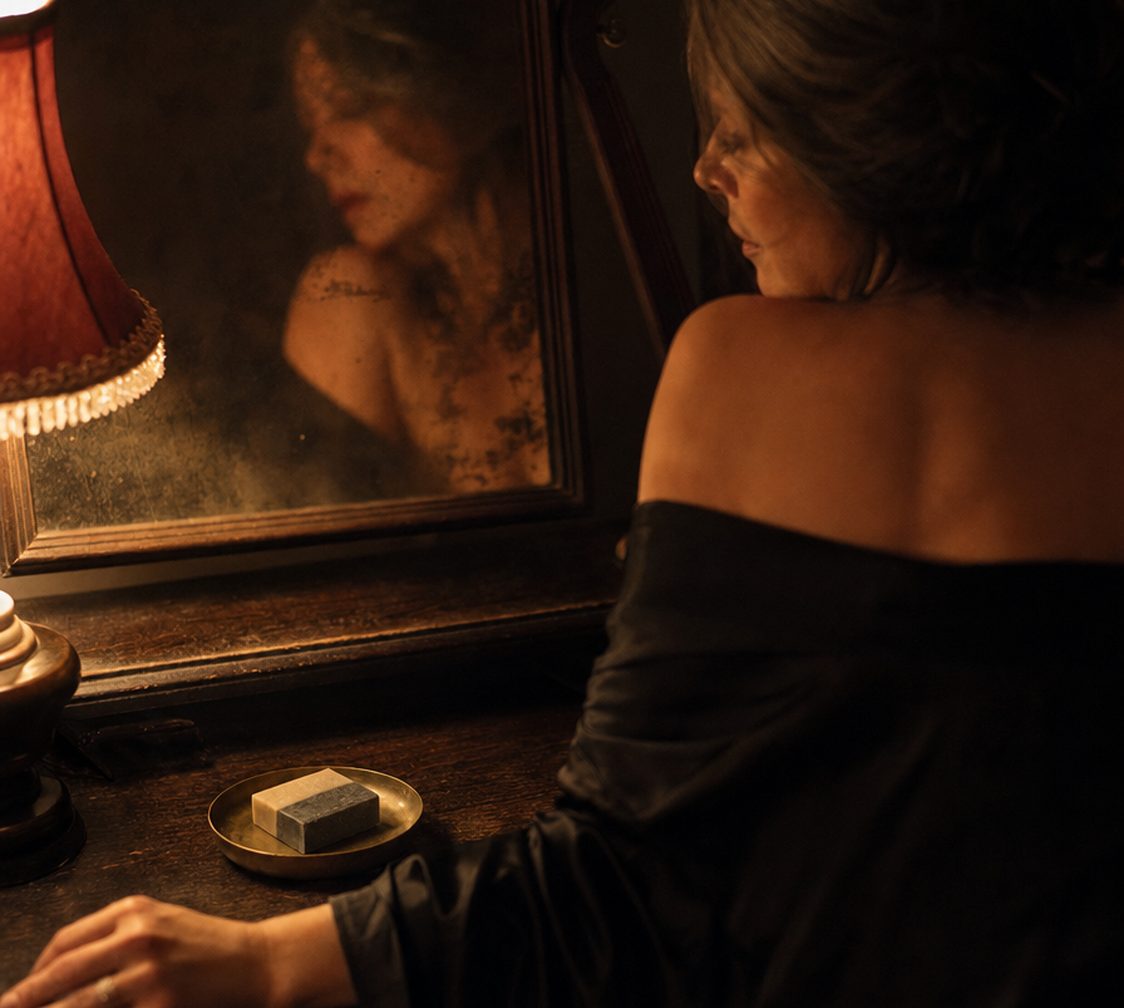 A woman at her mirror by a lamp at night, a bar of soap on a brass dish beside her