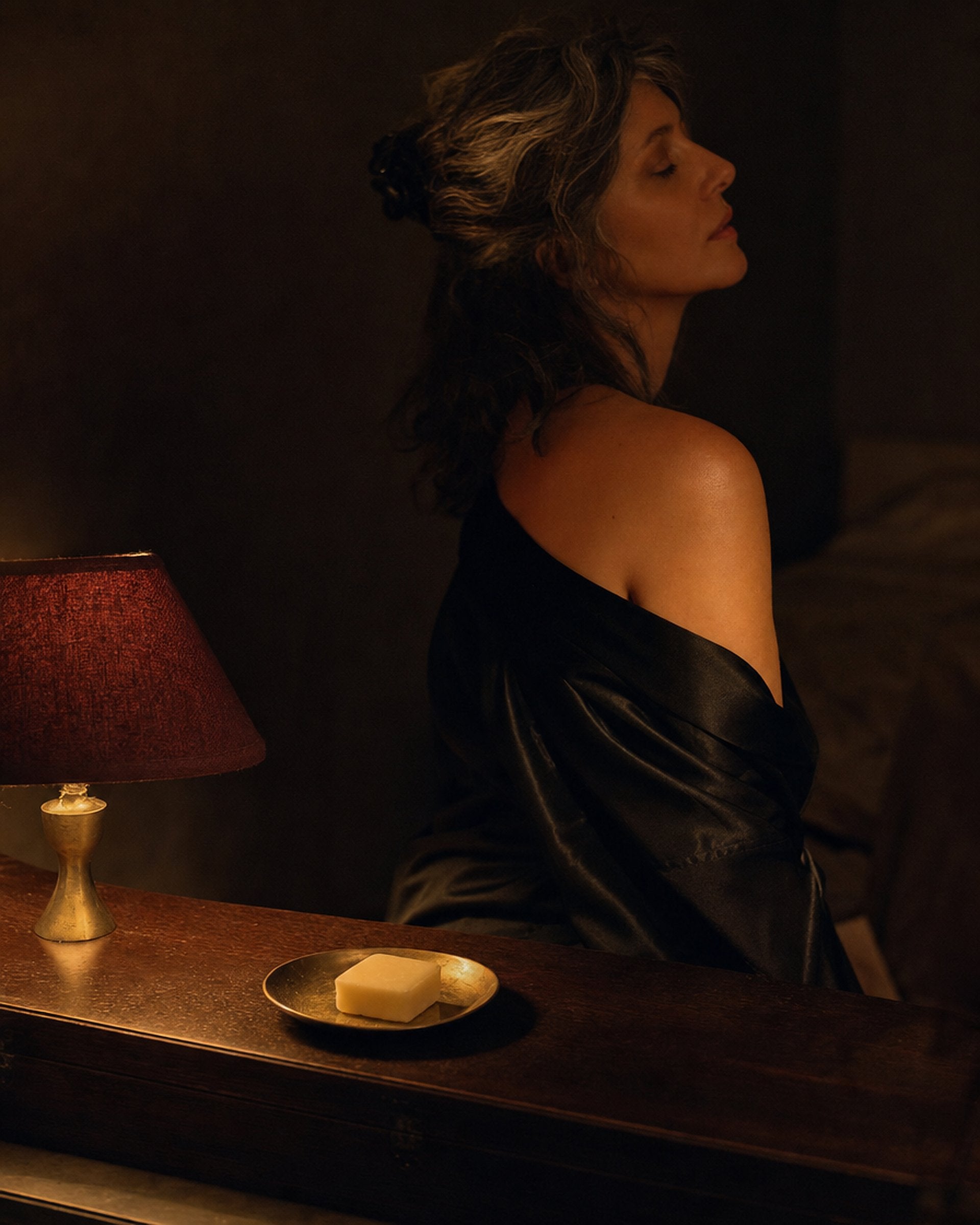 A woman in a black silk robe by a lamp at night, a bar of soap on a brass dish